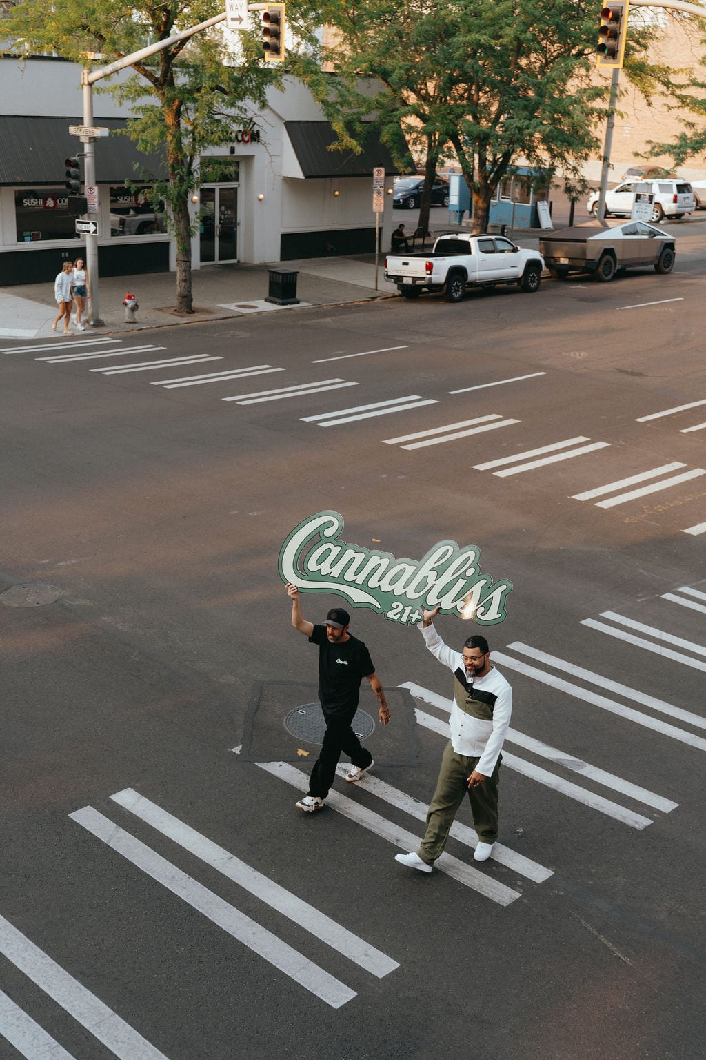 Two people carry the Cannabliss 21+ sign across a downtown crosswalk, seen from above.
